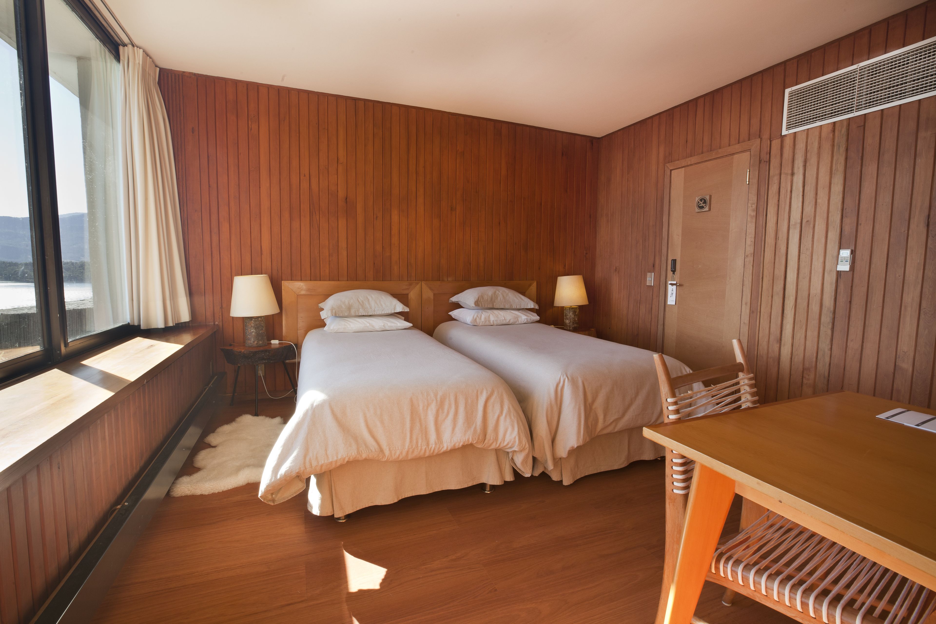 A room with two beds, wooden walls, a window with curtains, a table, and a chair.