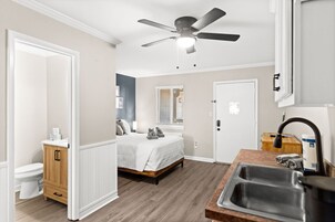 Habitación de hotel compacta con cama, baño pequeño, kitchenette y ventilador de techo.