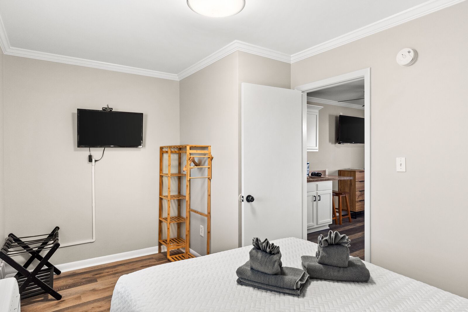 A bedroom with a bed, a TV mounted on the wall, a wooden shelf, and a bathroom visible through an open door.