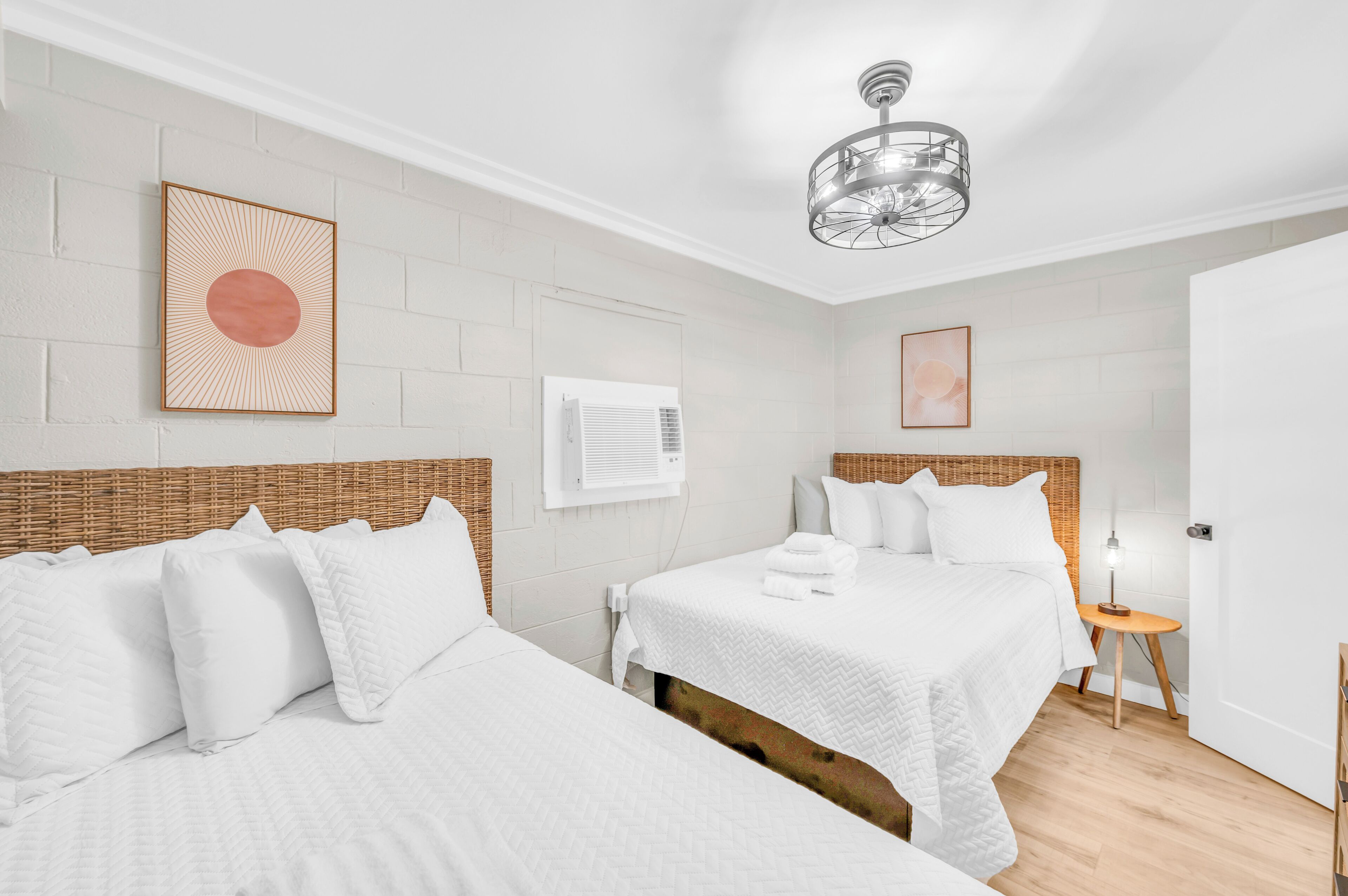 Habitación de hotel con dos camas, ropa blanca, cabecero de mimbre, una lámpara de araña moderna y una mesita de noche con lámpara.