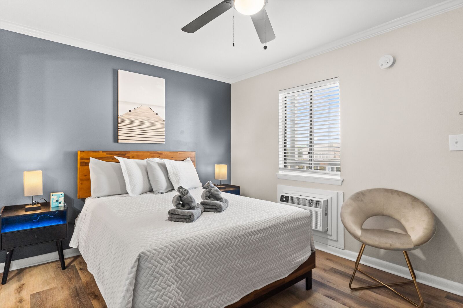 Une chambre moderne comprenant un lit, des tables de chevet, un ventilateur de plafond, une fenêtre avec des stores et une chaise.