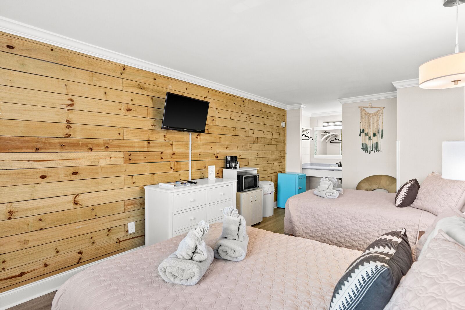 A hotel room with wooden panel walls, a flat-screen TV, a bed with a pink quilt, a bedside table with a coffee maker, and a small blue refrigerator.