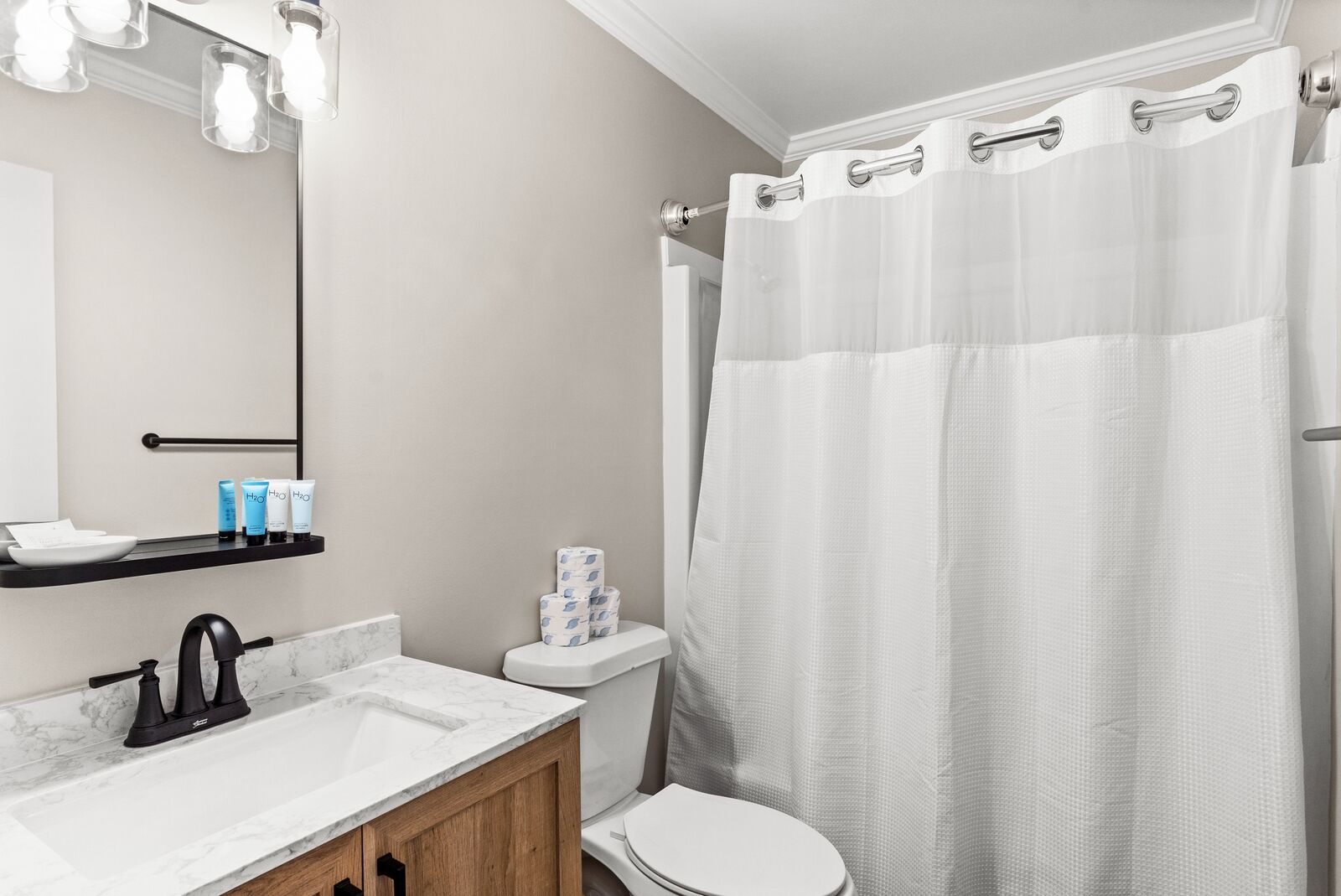 A bathroom with a white toilet, a sink with a marble countertop, a mirror with lights, and a shower curtain.