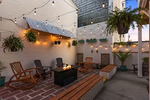 An outdoor patio with wooden chairs, a fire pit, string lights, and potted plants.