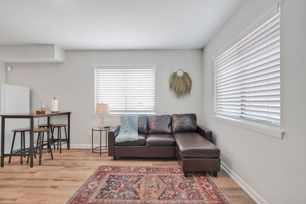 A modern living room with a brown leather sofa, a red patterned rug, a dining area with stools, and a small side table.