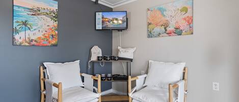 A corner room with two white cushioned chairs, a TV mounted on the wall, and two paintings depicting beach and coral scenes.