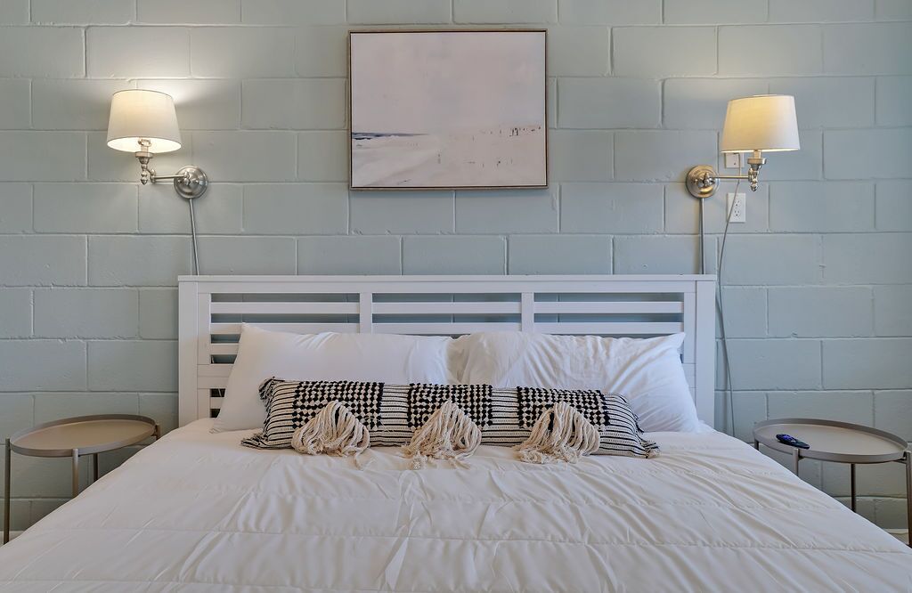 A neatly made bed with white bedding and decorative pillows, two bedside tables with lamps, and a framed artwork on the wall.