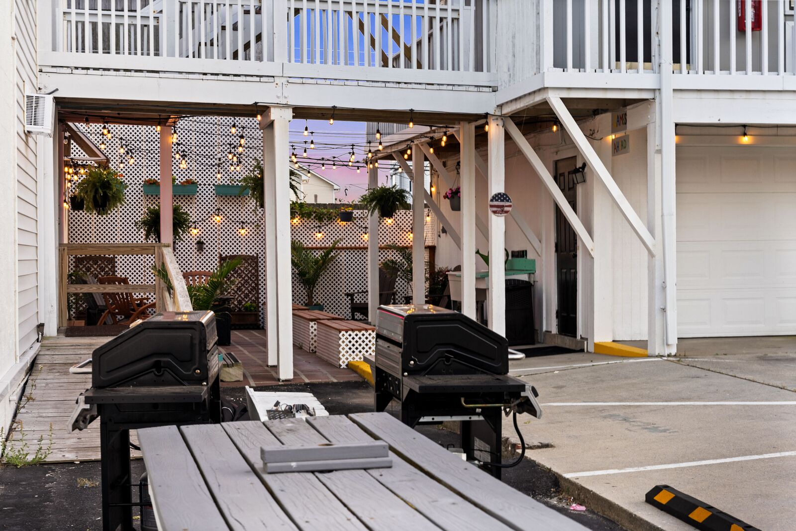Un patio al aire libre con mesas de madera y parrillas, un cerco blanco y luces colgantes.