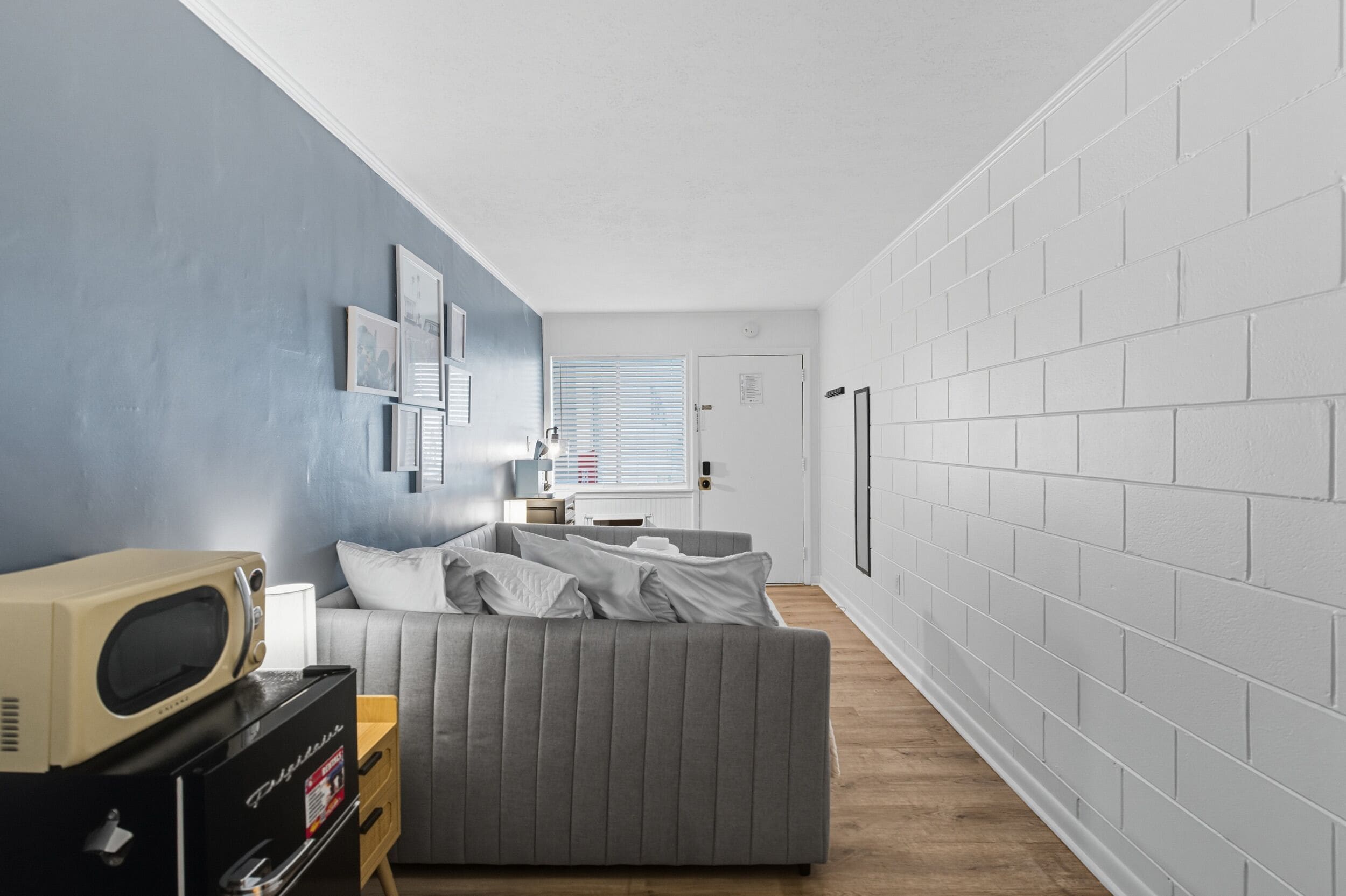 Ein kompakter Wohnbereich mit einem grauen Sofa, einer Mikrowelle und einem kleinen Holztisch. Eine weiße Tür und ein Fenster mit Jalousien sind zu sehen.