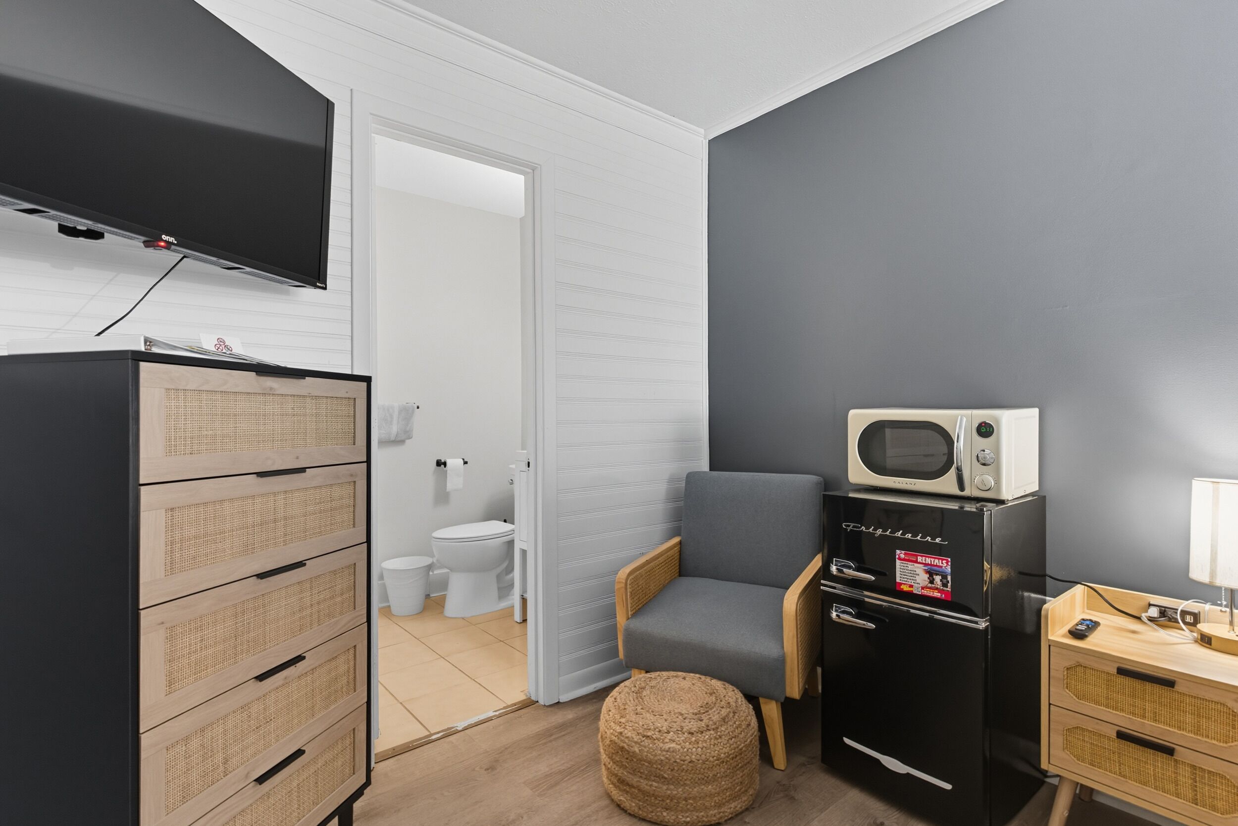 Una habitación compacta con televisión de pantalla plana, microondas, kitchenette pequeña y reposapiés de mimbre.