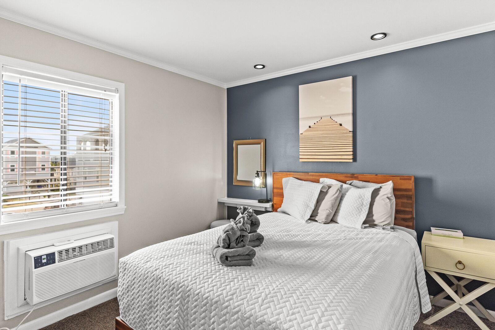 A bedroom with a bed, a nightstand, a wall-mounted mirror, and a window with blinds.