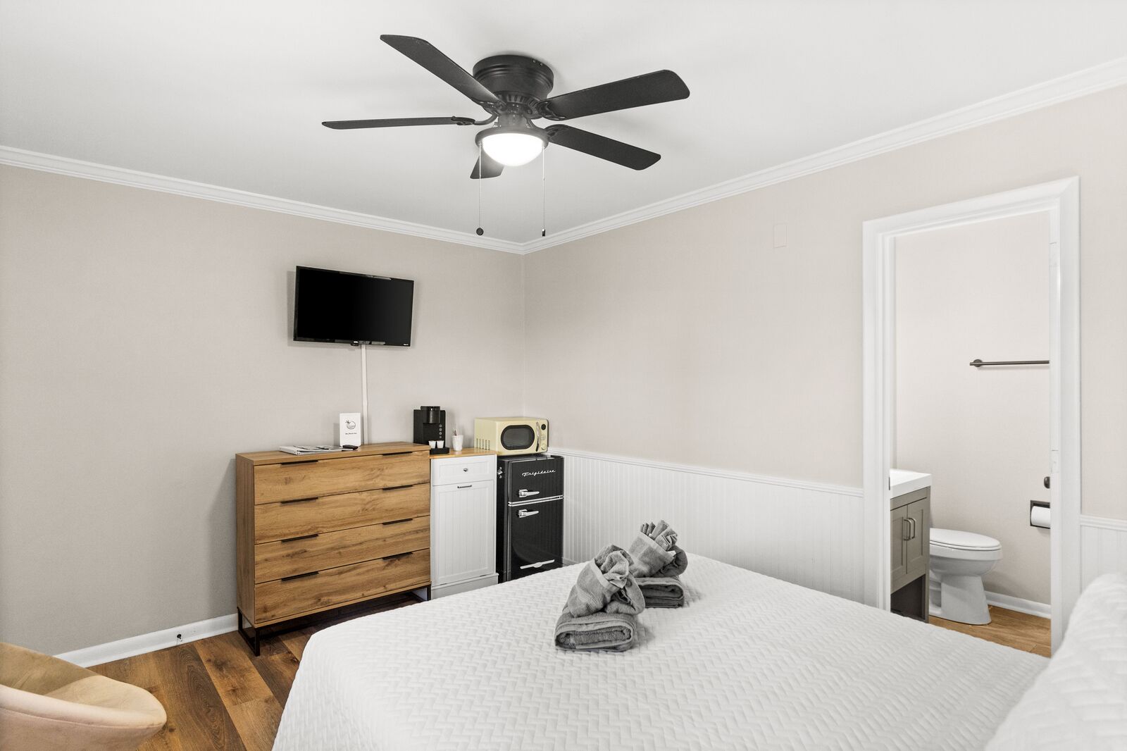 A bedroom with a bed, a wooden dresser, a ceiling fan, a television, a microwave, and a toilet.
