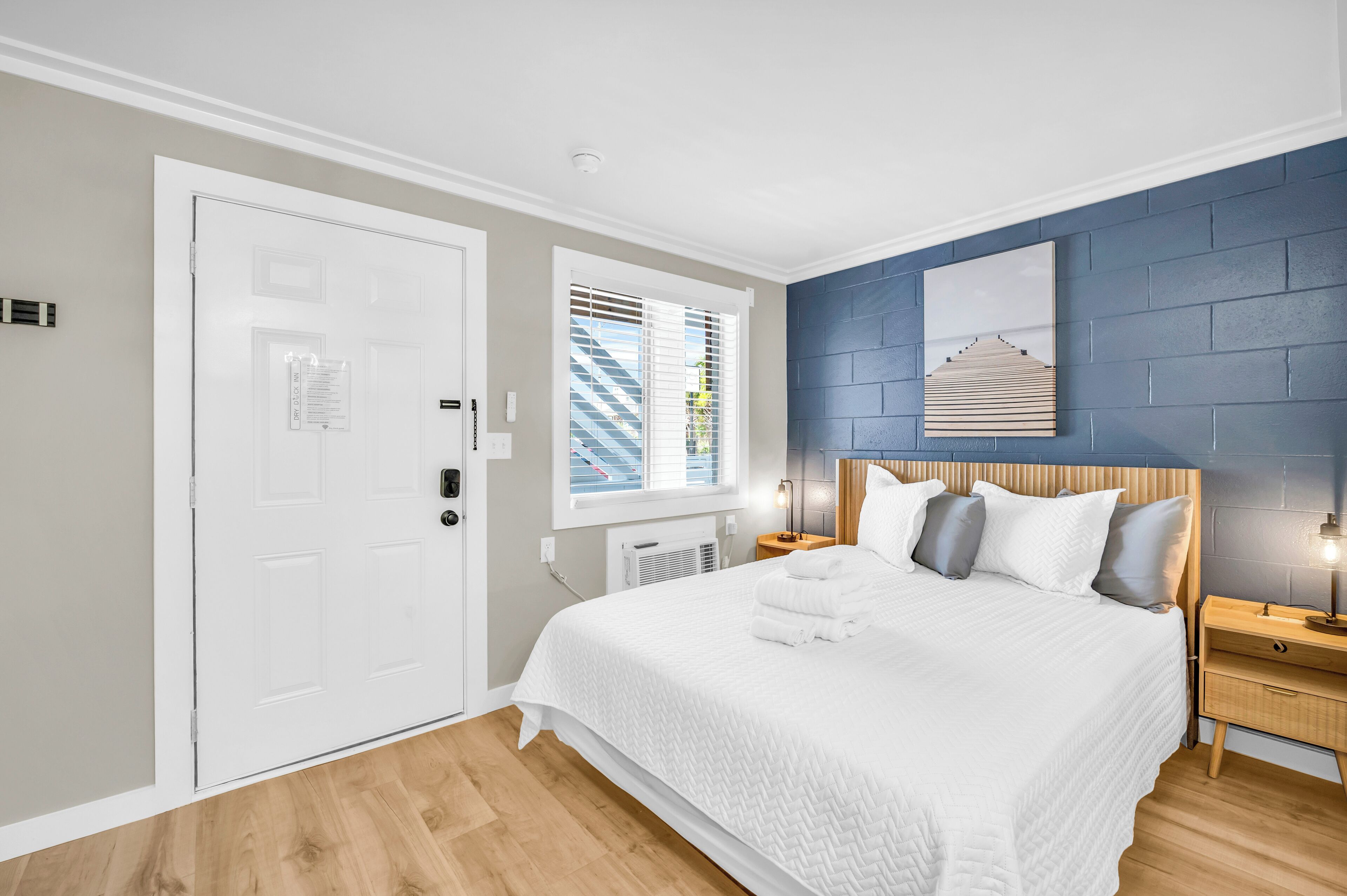 Habitación de hotel con una cama, una mesita de noche, una lámpara de pared, una ventana con persianas y una puerta.