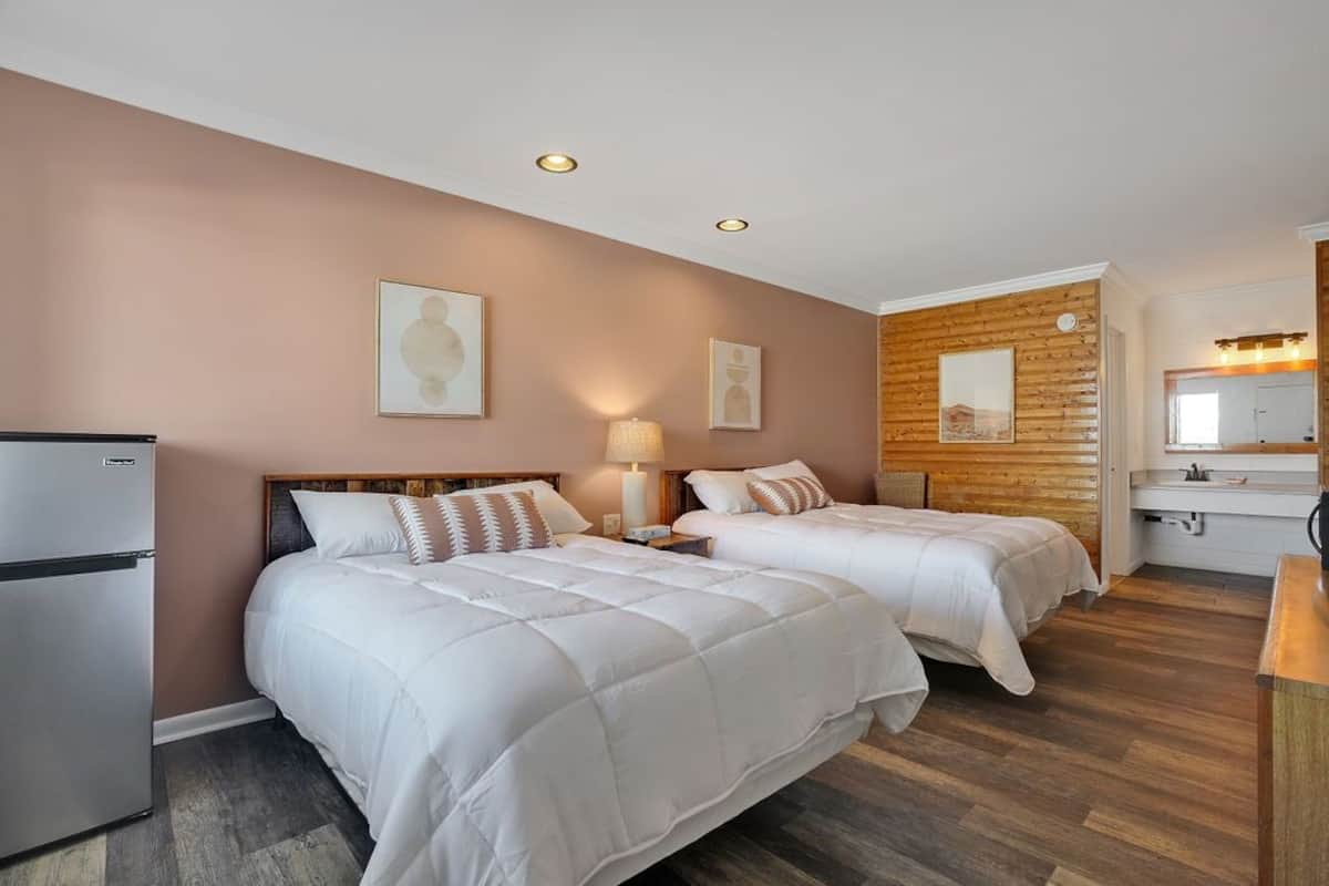 A hotel room with two beds, a refrigerator, a wooden accent wall, and a bathroom with a mirror and sink.