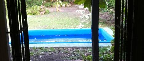Une porte ouverte donne sur un patio avec une piscine bleue, entouré de verdure et doté d’un sol carrelé.