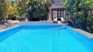 A rectangular swimming pool with a clear blue water surface, surrounded by steps leading into the pool. There is a covered seating area with two lounge chairs and a table. - Hacienda San Francisco (Dzidzantun)