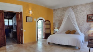 A bedroom with a bed under a white canopy, a wooden door, a small table with a lamp, and a wall-mounted light fixture. - Hacienda San Francisco (Dzidzantun)