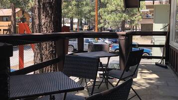 A covered outdoor seating area with wicker chairs and tables, overlooking a parking area and buildings.