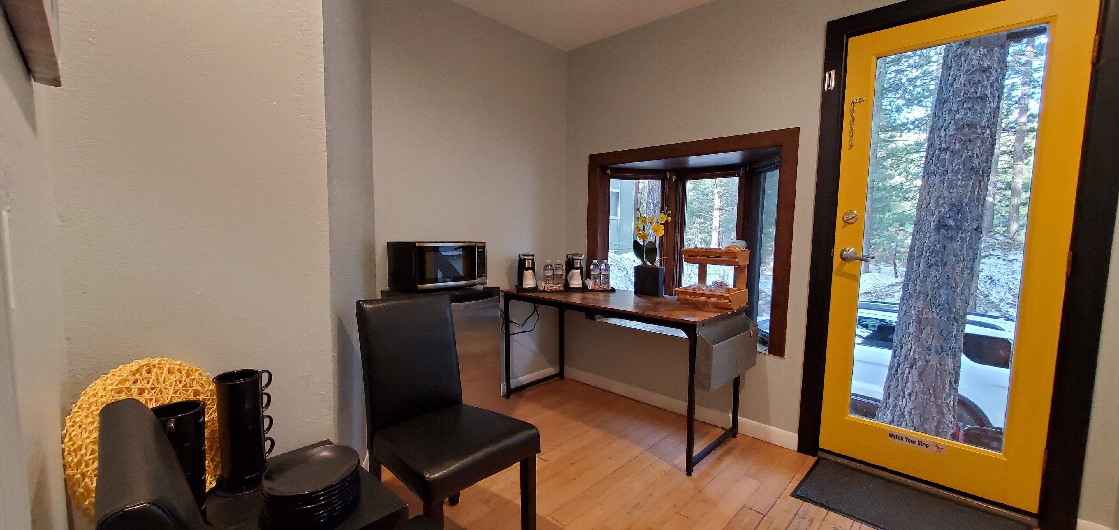 Habitación pequeña con piso de madera, una silla negra, una mesita con televisor, una ventana que da a un árbol y un coche, y una puerta con letrero que dice 'Precaución, Escalón'.