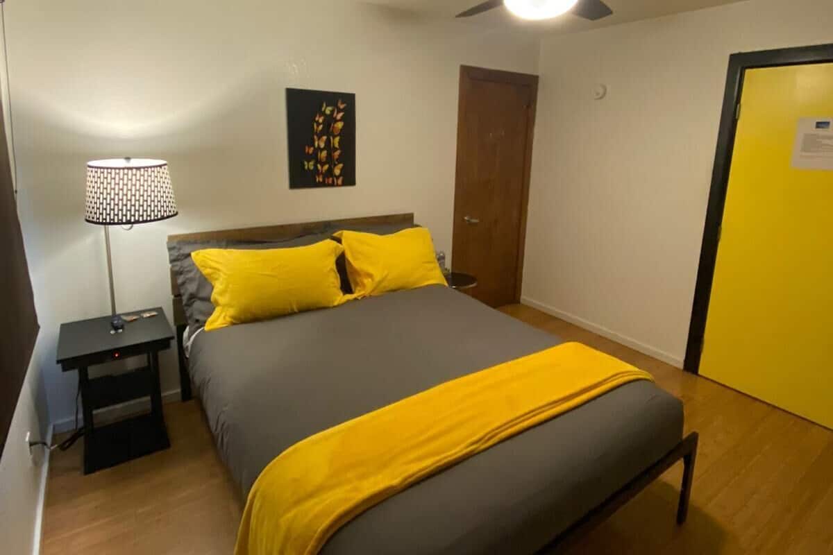 A bedroom with a bed, a ceiling fan, a nightstand, a lamp, a door, and a wall with a picture.