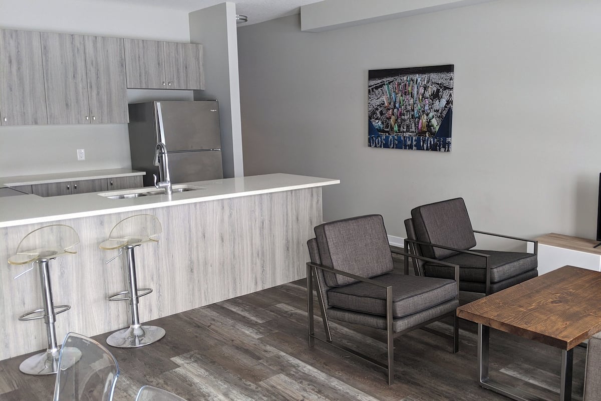 A modern kitchen with a built-in refrigerator, a bar area with stools, a seating area with chairs, a wooden coffee table, and a flat-screen TV.
