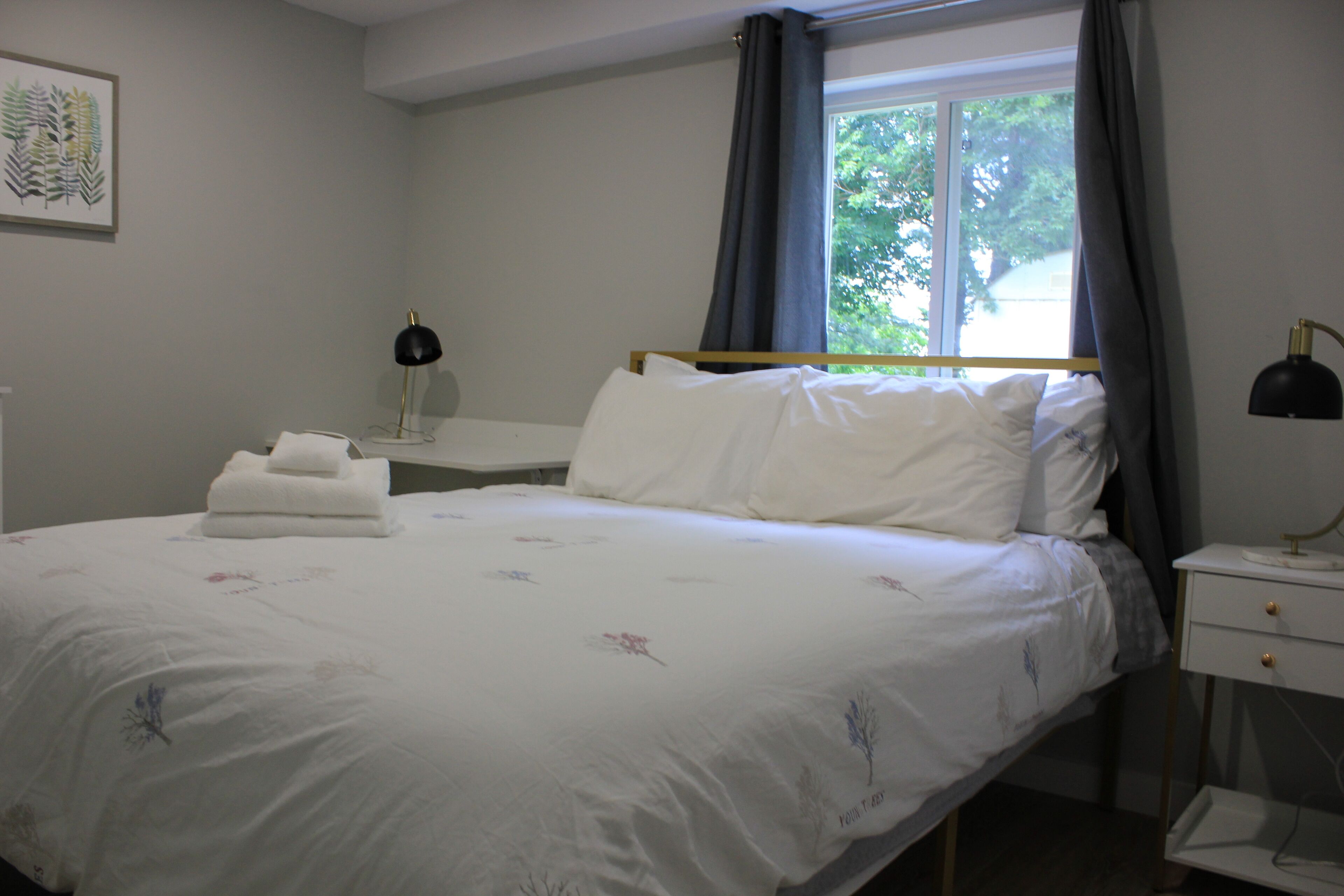 A neatly made bed with white linens and floral patterns, a bedside table with a lamp, and a window with curtains.