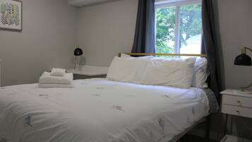 A neatly made bed with white linens and floral patterns, a bedside table with a lamp, and a window with curtains.