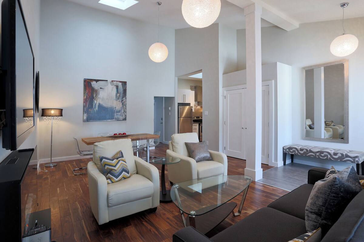 Una sala de estar moderna con un sofá, sillones, una mesa de centro de vidrio y un televisor. Hay un comedor con una mesa y sillas, y una cocina al fondo.
