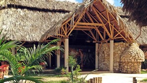 Estructura de techo de paja con pared de piedra, terraza de madera y plantas tropicales.