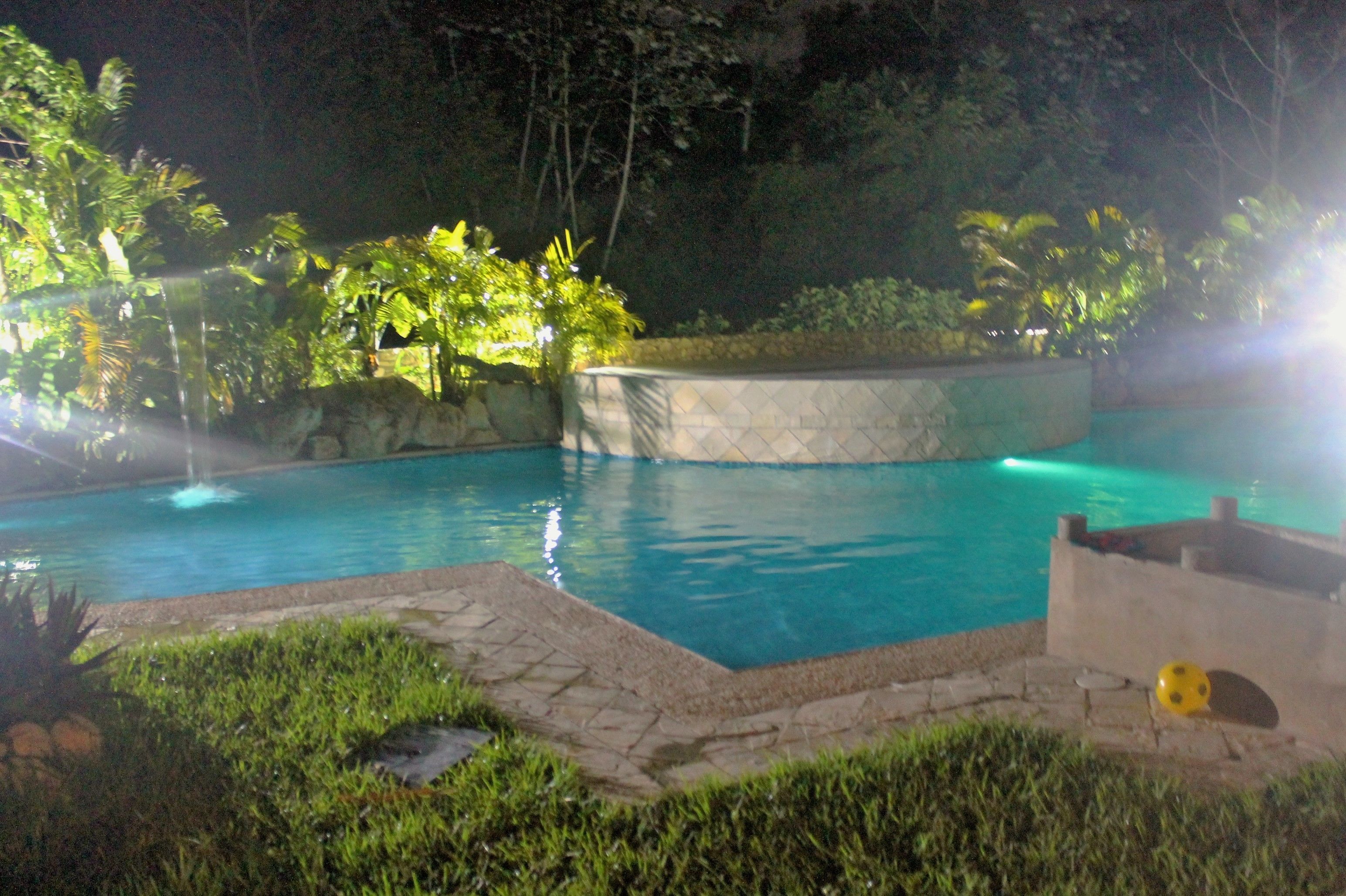 A nighttime view of a swimming pool with a fountain, surrounded by lush greenery and a stone wall.