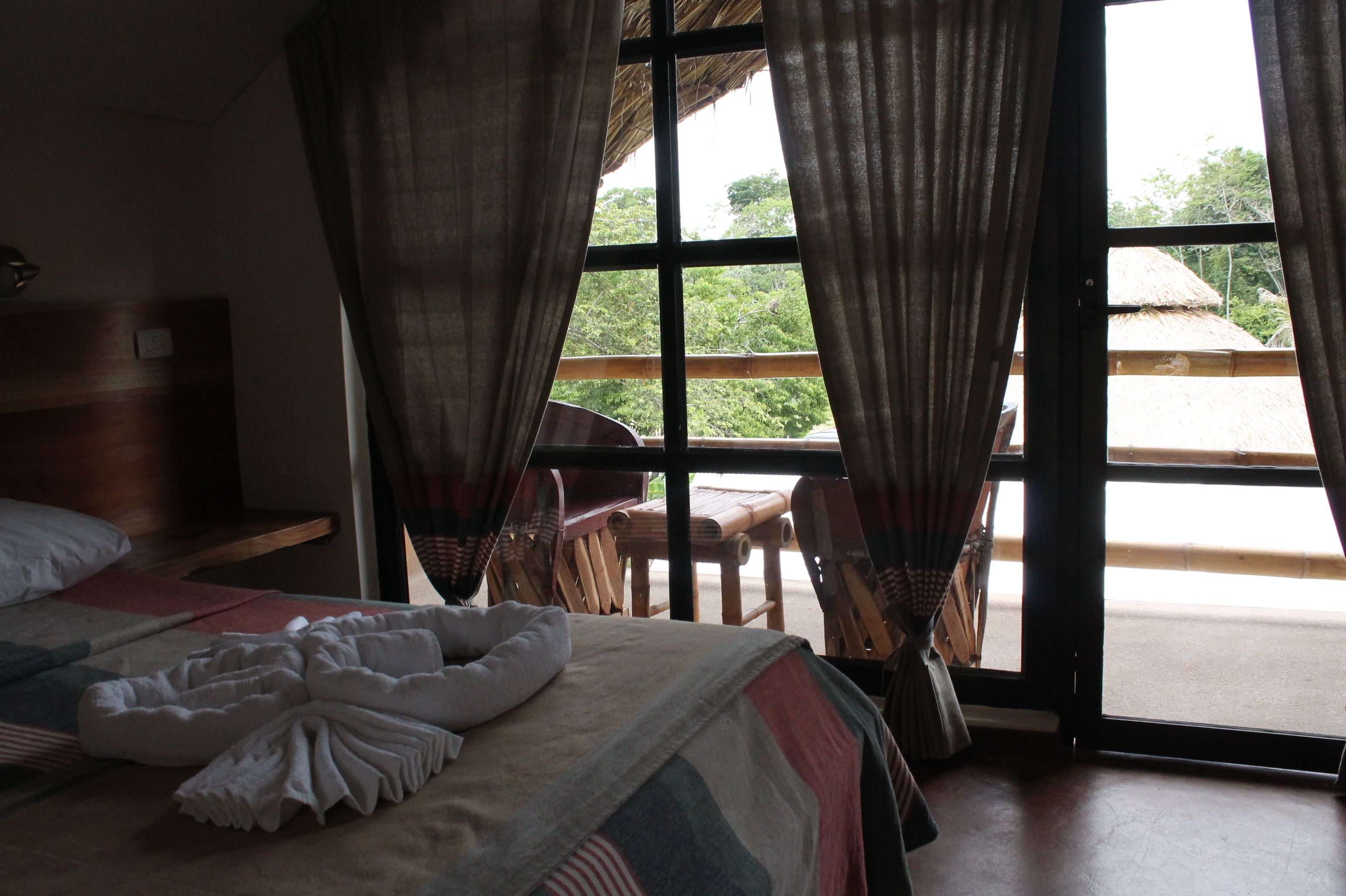 Ein Hotelzimmer mit Bett, Holz-Kopfteil, einem Fenster mit Vorhängen und Blick ins Freie.