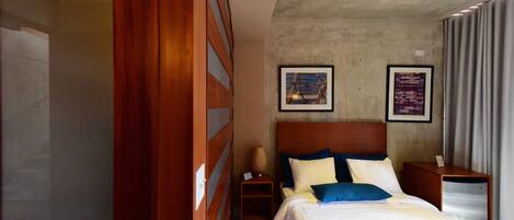 Habitación de hotel con cama, cabecera de madera, dos cuadros en la pared, una mesita de noche y una pequeña mesa.