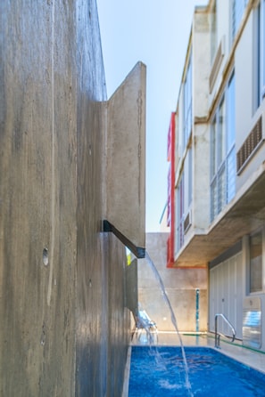 A swimming pool with a concrete structure, a water fountain, and a building with balconies in the background.