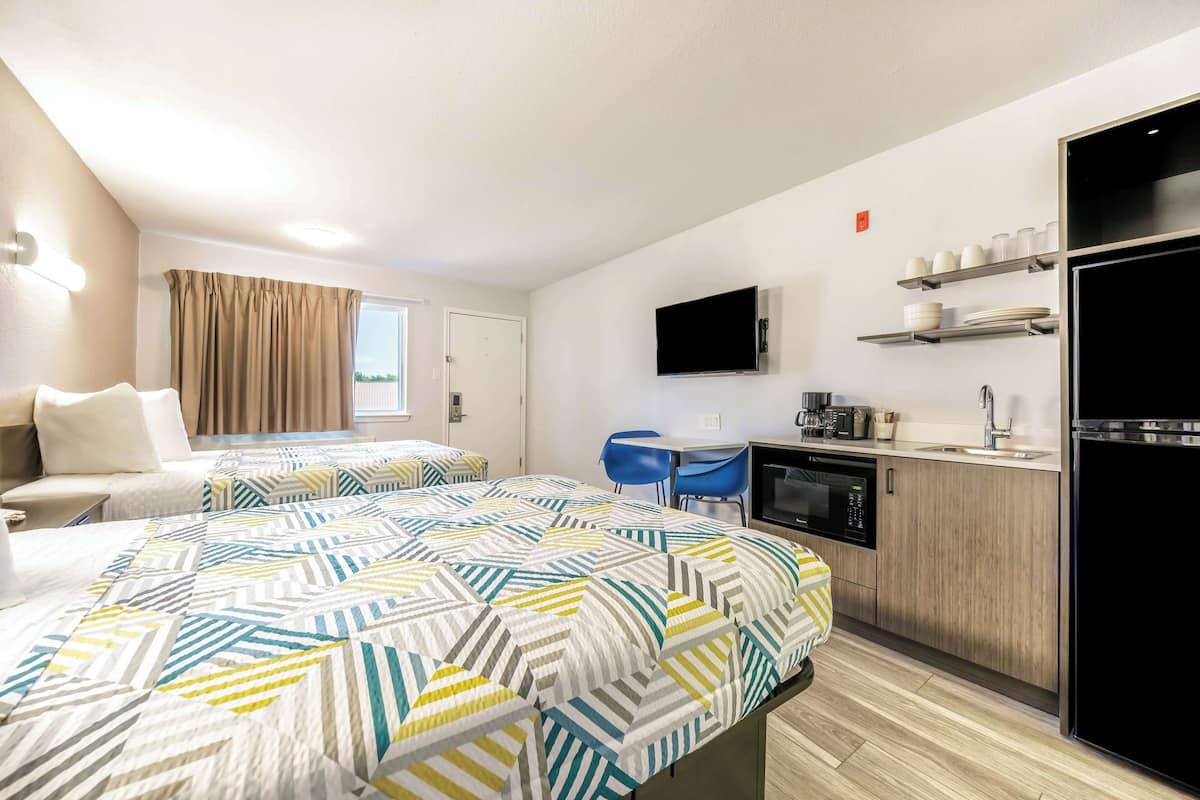 A modern hotel room with a large bed, a desk, a kitchenette, and a TV.