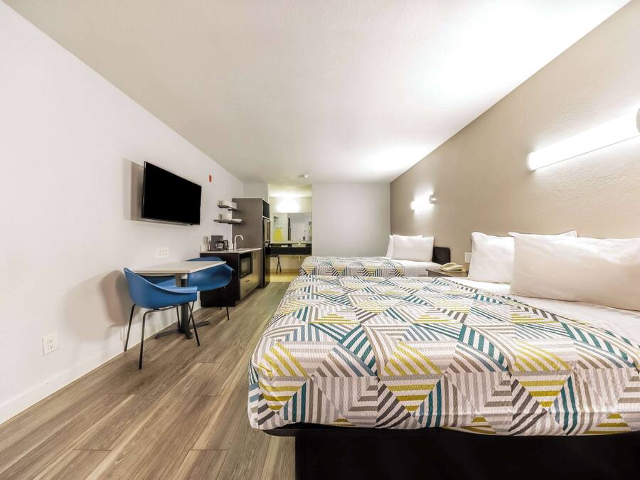 A hotel room with a bed, a desk, a chair, a TV, and a kitchenette.