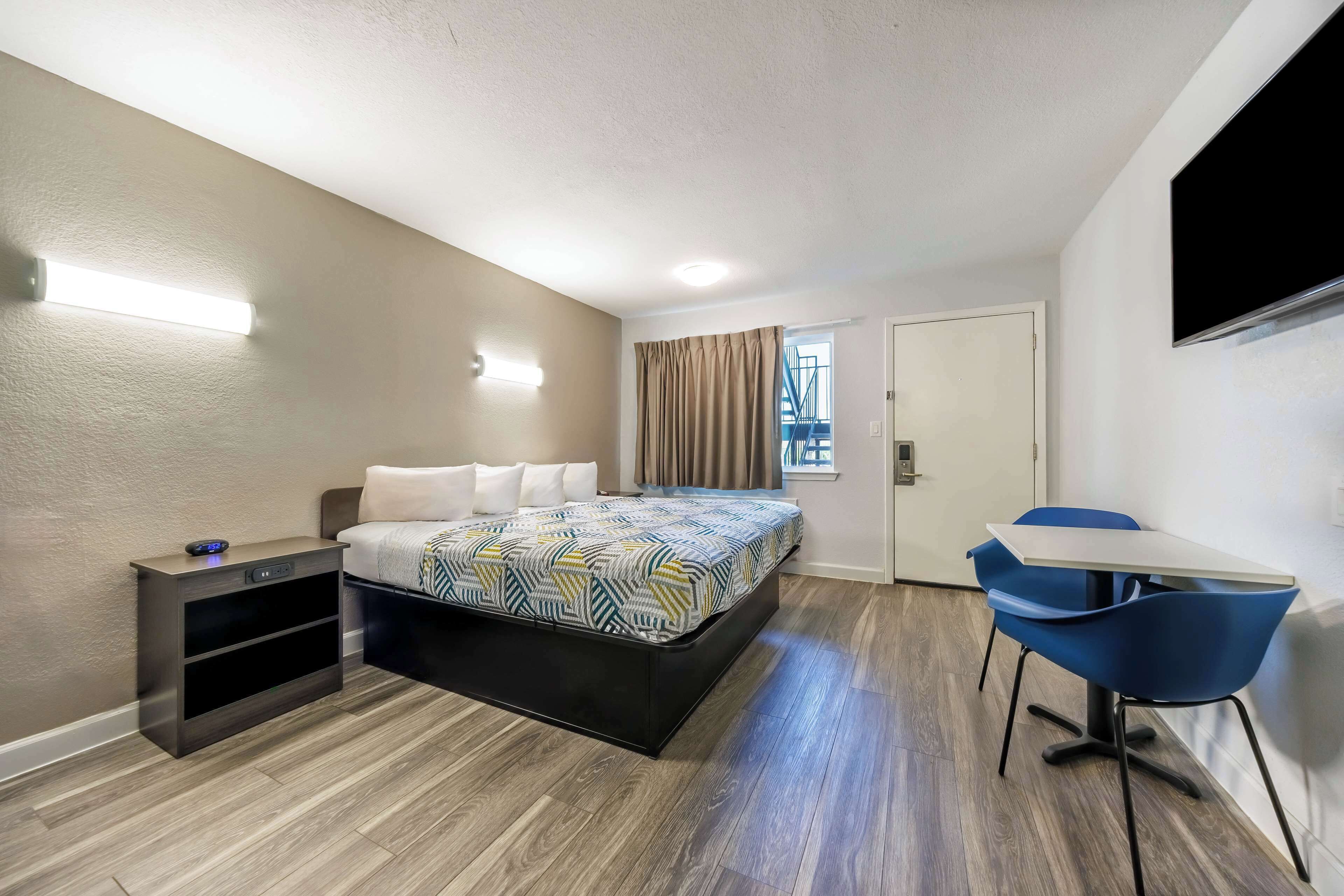 A compact hotel room with a bed, bedside table, chair, and a small desk.