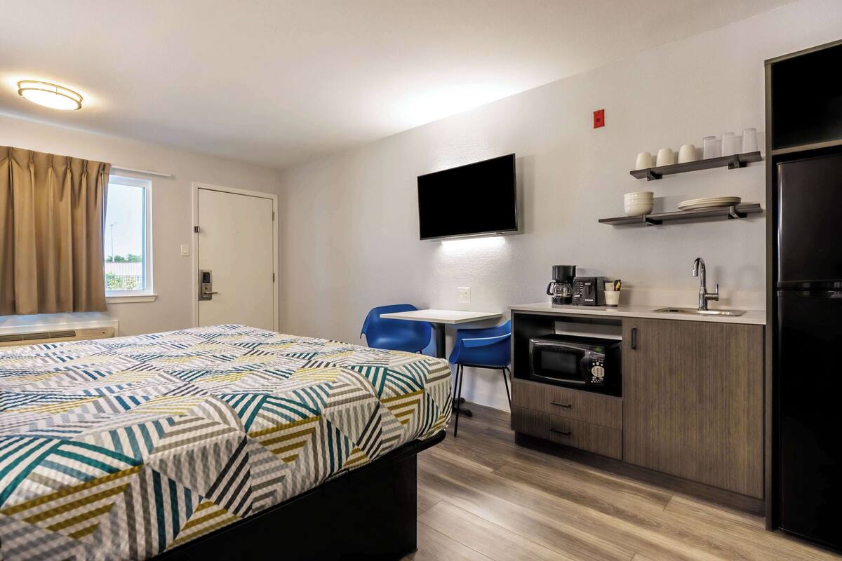 A modern hotel room with a bed, a desk, a flat-screen TV, a microwave, a sink, and a refrigerator.