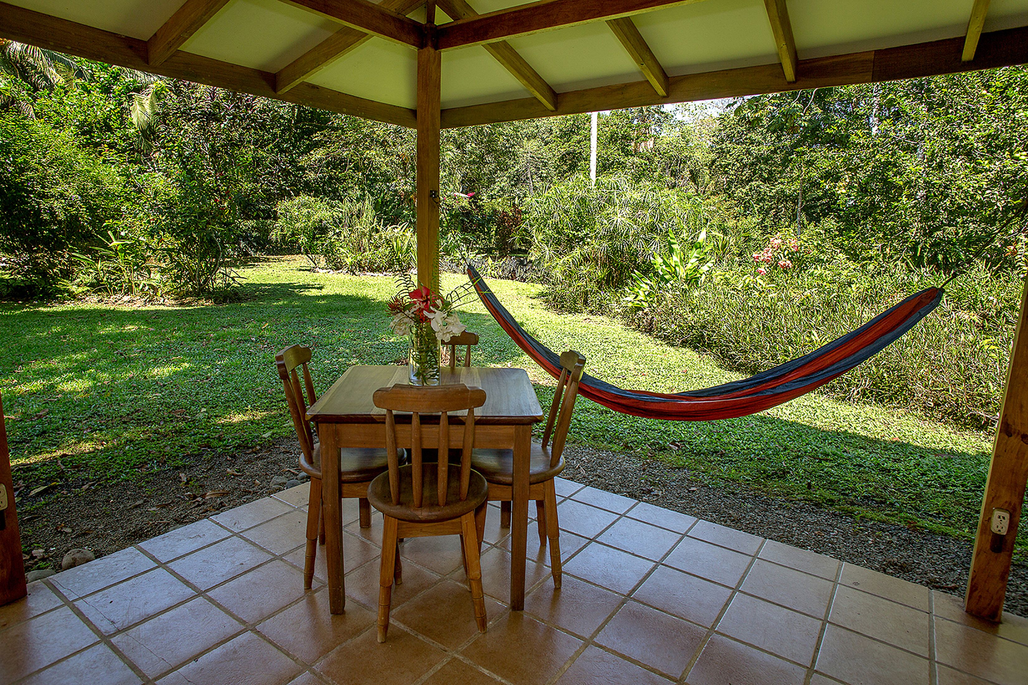 Une terrasse couverte avec une table et des chaises, un hamac et une vue sur un jardin luxuriant.