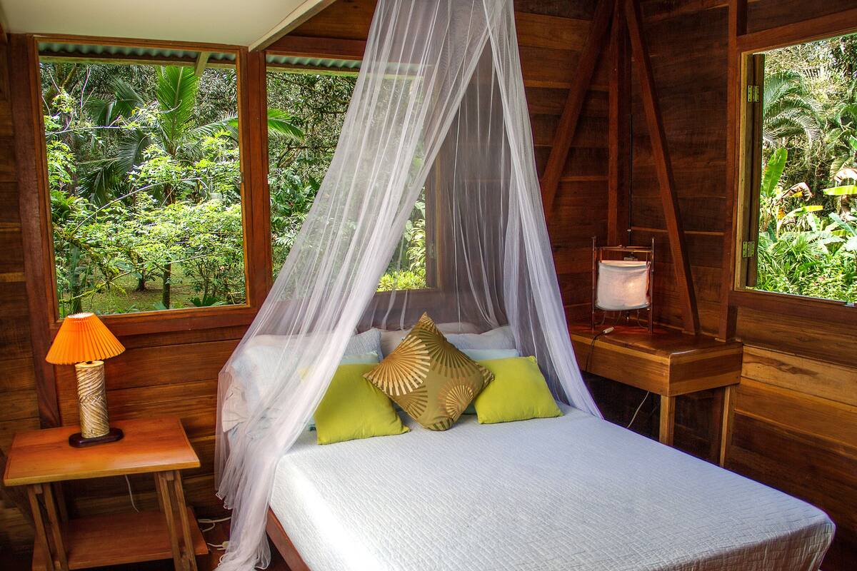 Ein hölzernes Himmelbett mit weißer Bettdecke und vielen Kissen, ein Nachttisch mit Lampe sowie Blick auf üppiges Grün durch große Fenster.