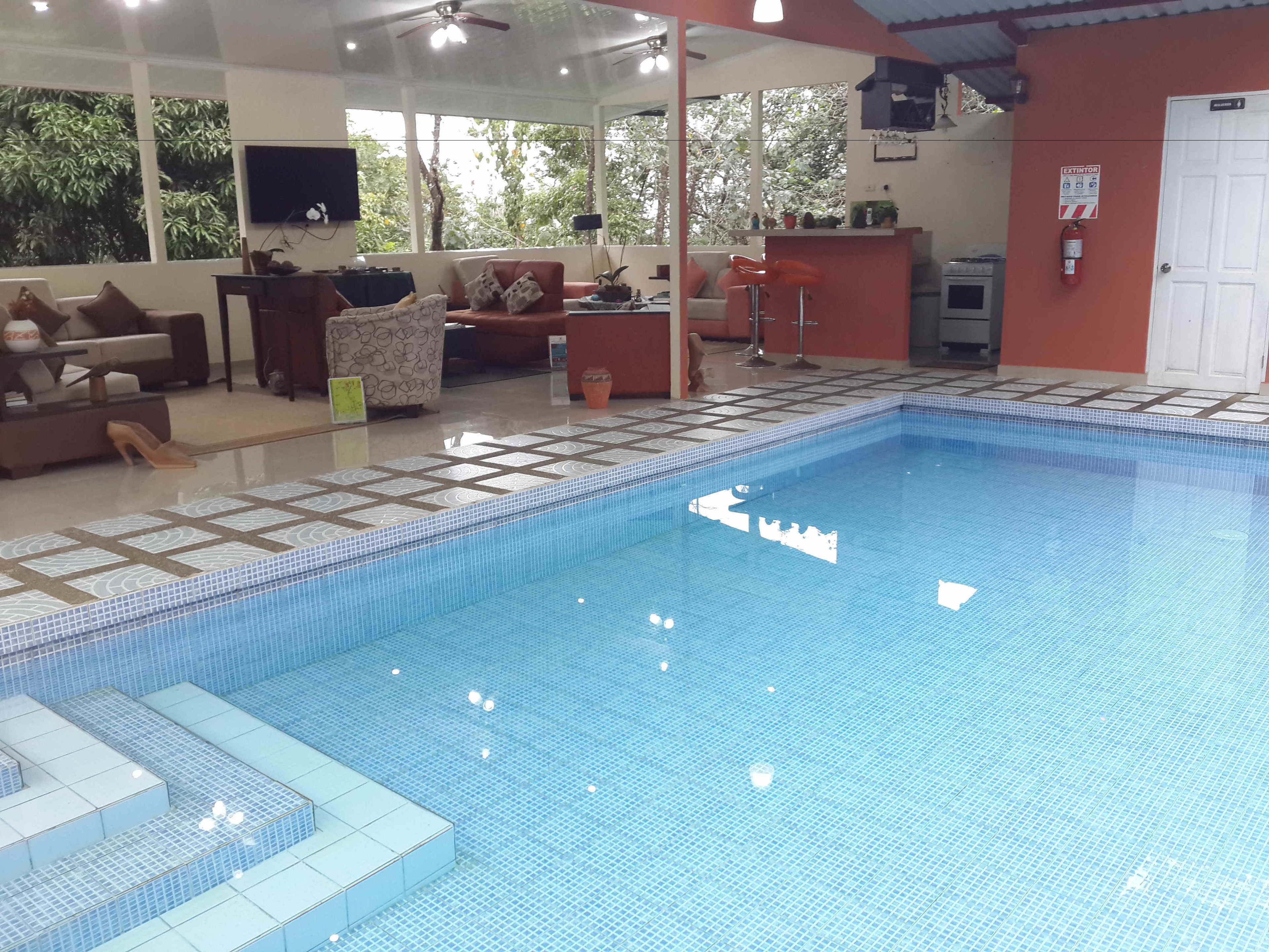 Une piscine avec des marches carrelées, un canapé, une télévision et un ventilateur de plafond dans une pièce donnant sur la forêt.