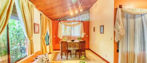 A room with a wooden ceiling, a dining table with chairs, a sofa, and a window with curtains.