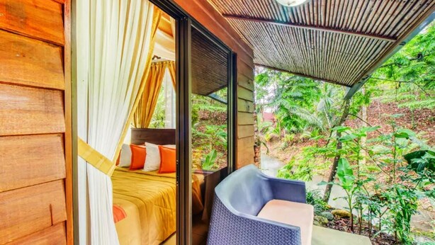 A wooden room with a bed, a chair, and a view of a lush garden. - San Rafael Ecolodge (La Tigra)