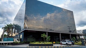 Een modern gebouw met een spiegelende glazen gevel, een plat dak en een parkeerplaats met verschillende voertuigen.