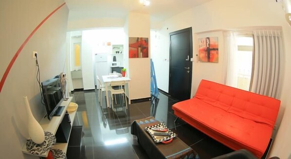 A compact living space with a red sofa, a dining table, and a kitchen area in the background.