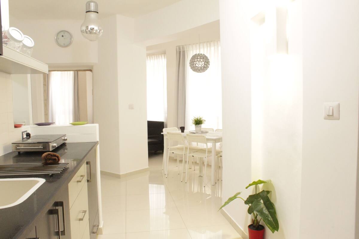 A modern kitchen with a black countertop, white cabinets, and a built-in oven. There is a dining area with a white table and chairs, and a potted plant on the side.