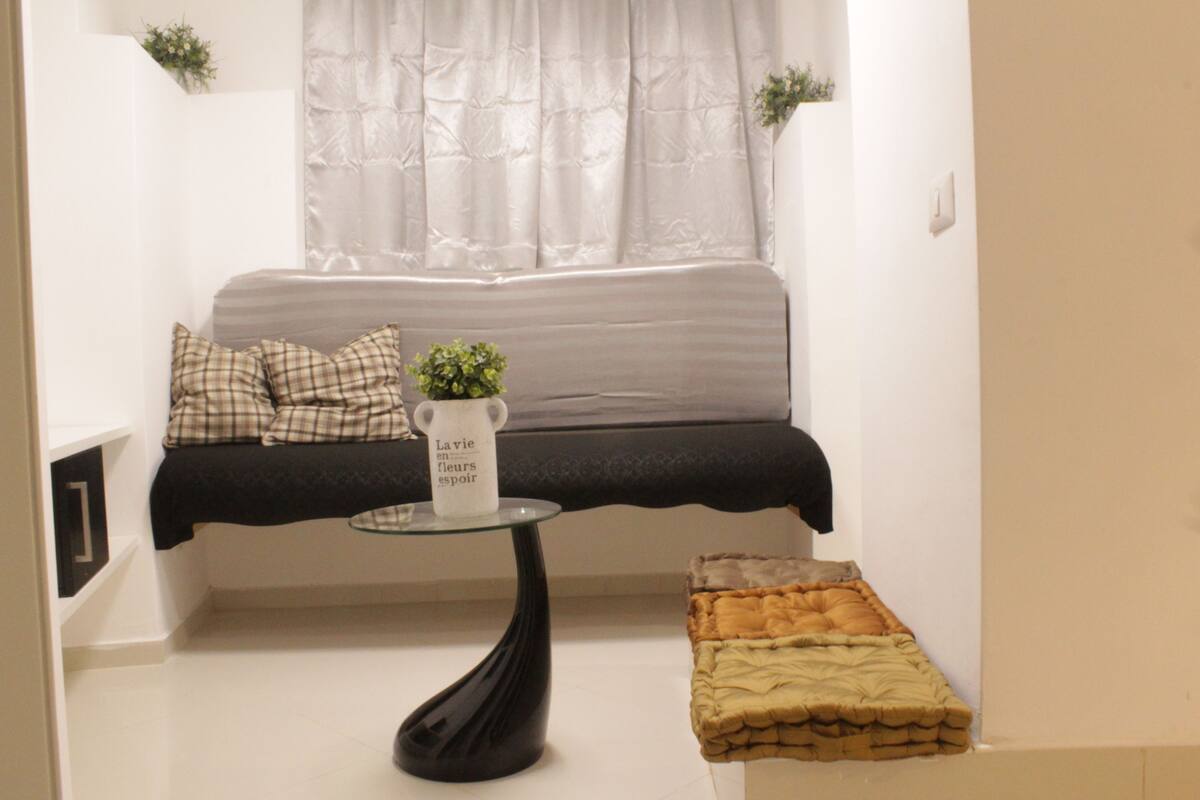 A small, cozy room with a bed, a glass table with a vase, and a bench.