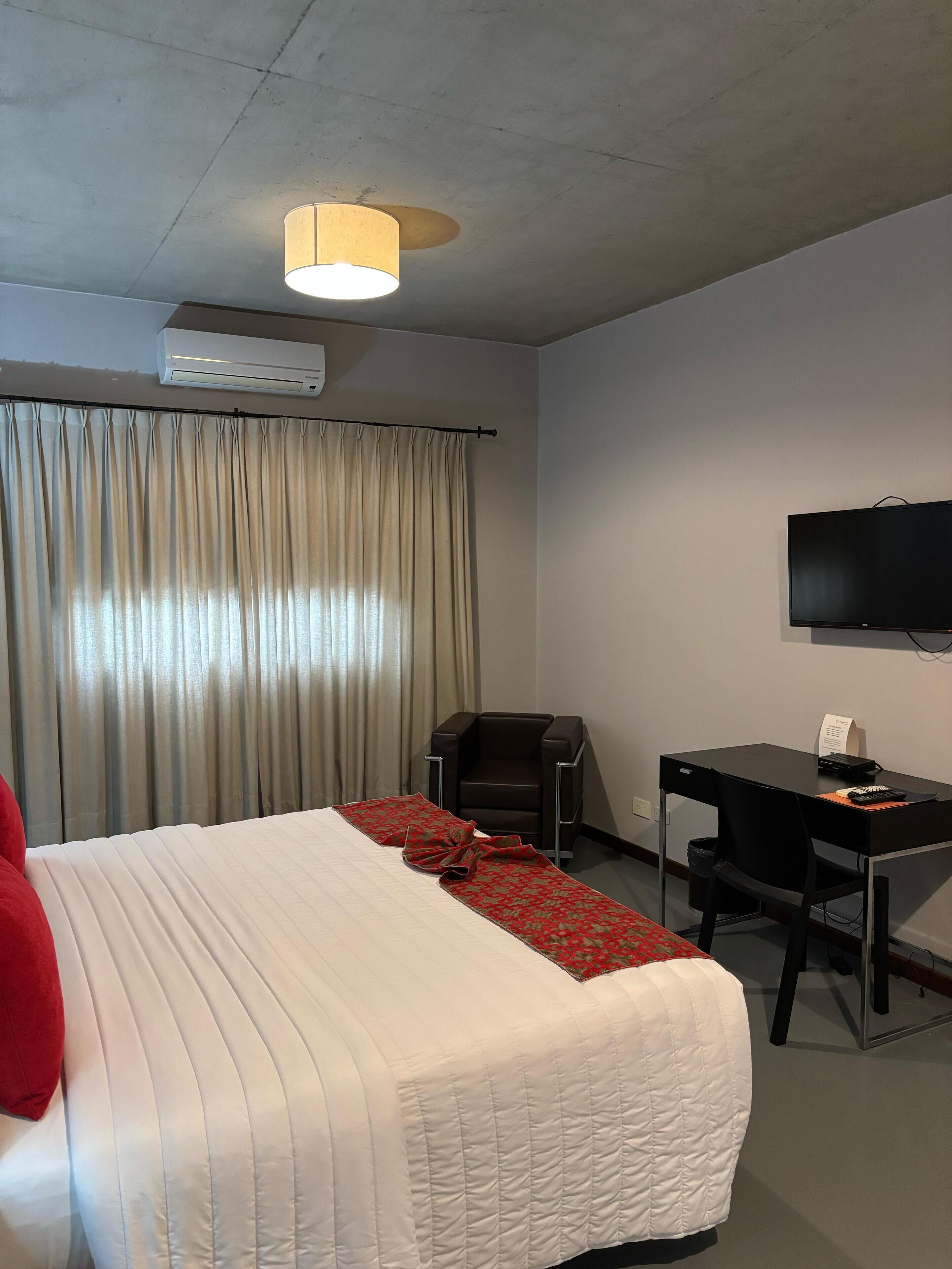 Habitación de hotel con cama, escritorio, silla, televisor y aire acondicionado.