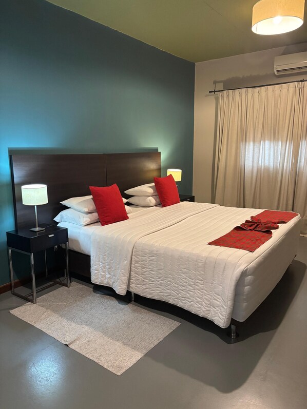 A neatly made bed with white bedding and red pillows, a bedside table with a lamp, and a large window with curtains.
