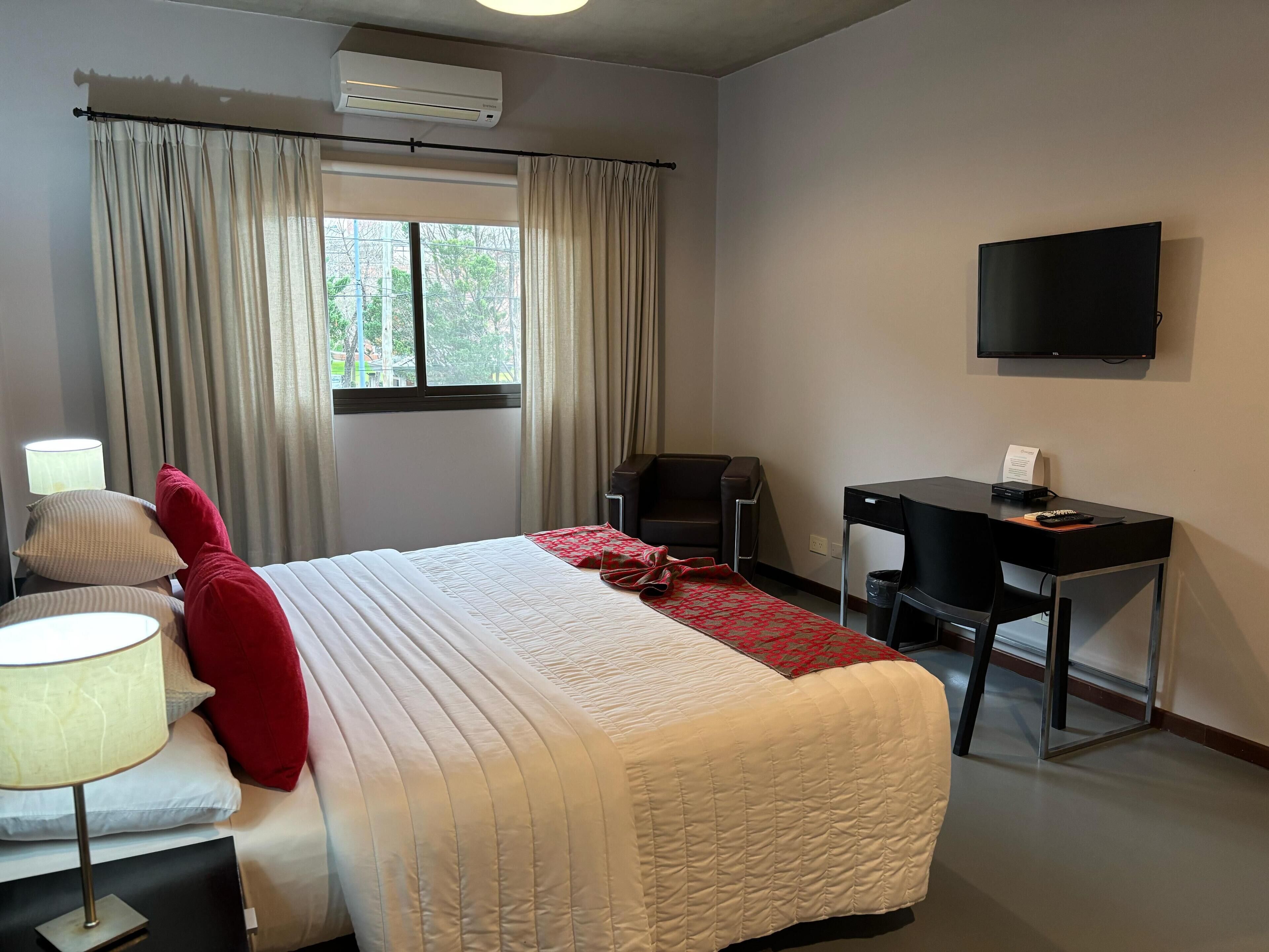 A hotel room with a bed, a desk, a chair, a TV, and an air conditioning unit.