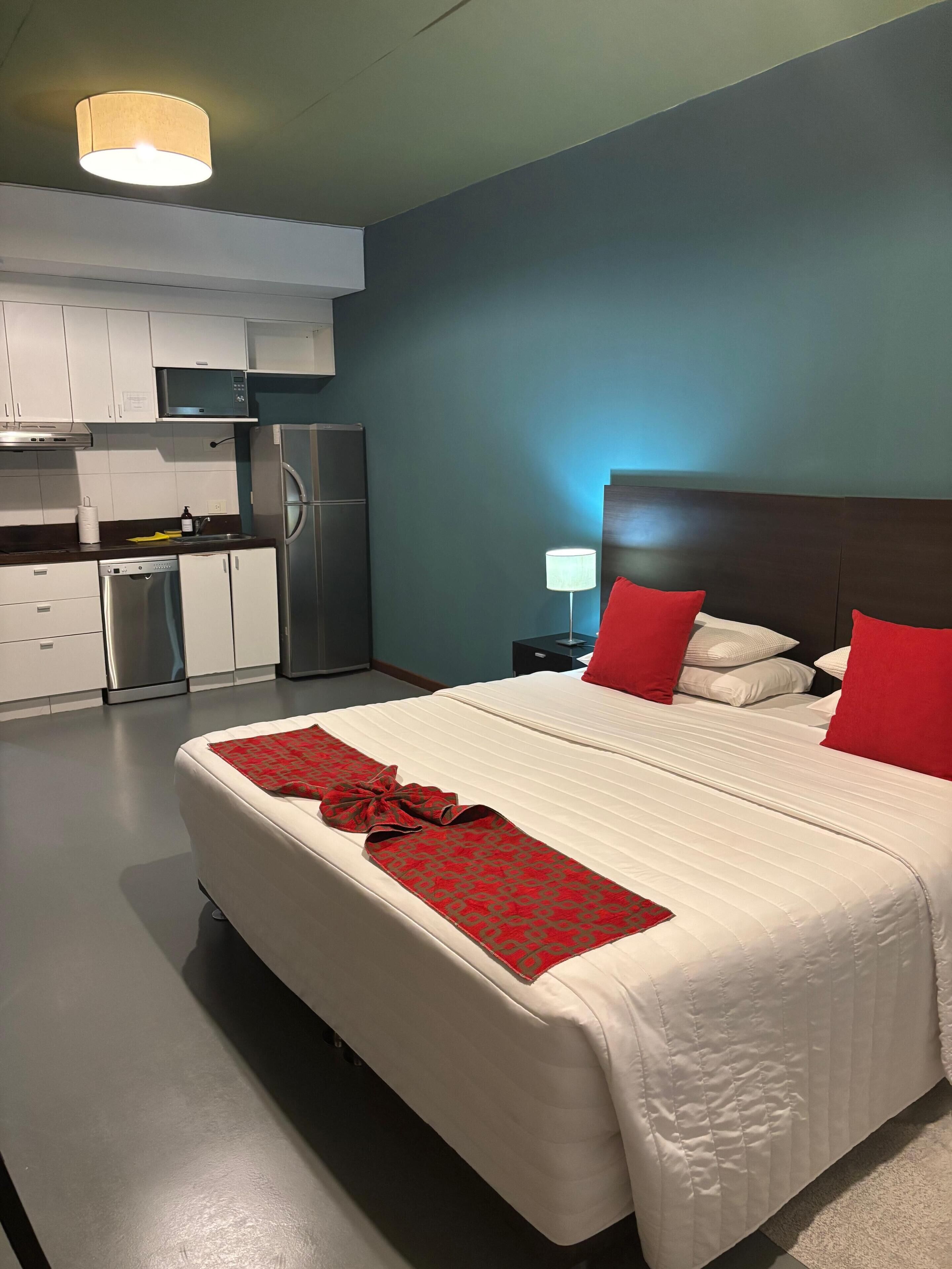 A hotel room with a bed, a nightstand, a lamp, a red throw blanket, and a kitchenette with a refrigerator and stove.